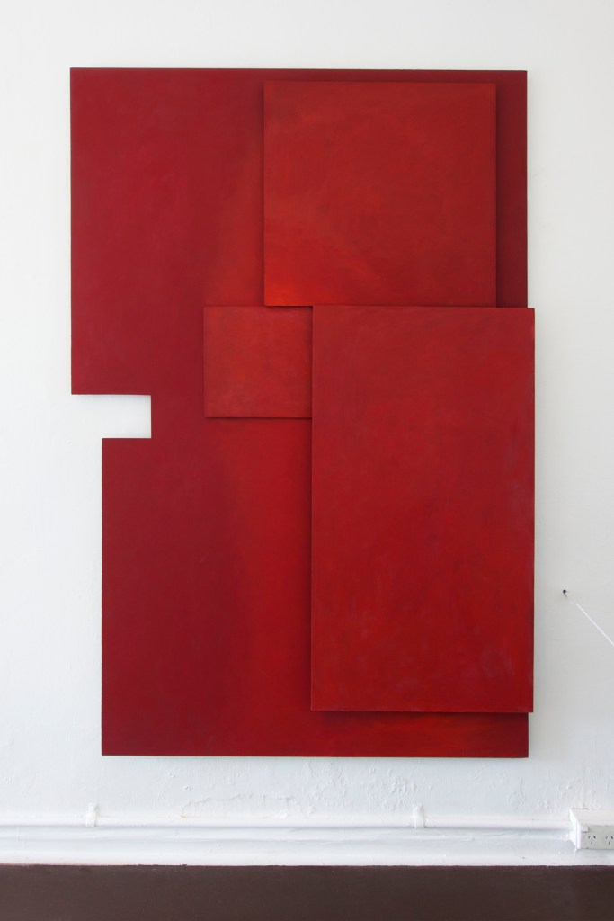 Documentation from the contemporary art installation In the Year Twenty Twenty One by Alex Gawronski held at KNULP gallery in Camperdown, Sydney, Australia. A red hard-edged three dimensional painting made up of different size rectilinear panels against a white wall above a brown floor.