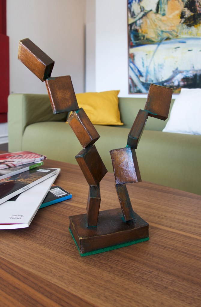 Documentation from the contemporary art installation In the Year Twenty Twenty One by Alex Gawronski held at KNULP gallery in Camperdown, Sydney, Australia. A close up of what appears to be a bronze abstract sculpture in the shape of small boxes stacked diagonally end on end on a brown wood grain coffee table. On the left, the corner of a stack of contemporary art magazines. An olive colored couch with a yellow and white cushion and an expressive abstract painting on a white wall.
