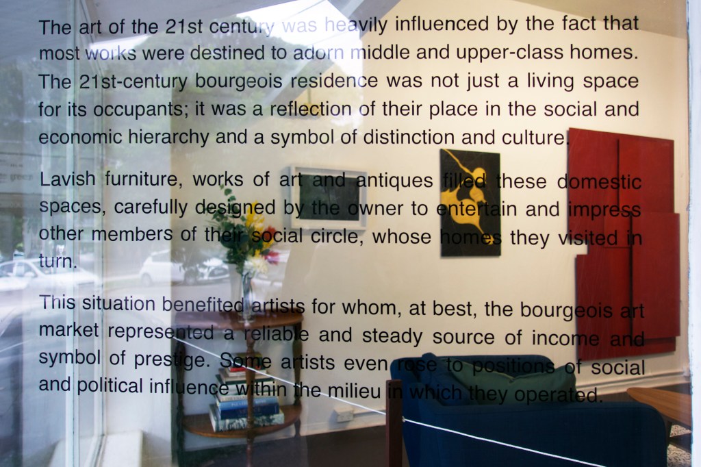 Documentation from the contemporary art installation In the Year Twenty Twenty One by Alex Gawronski held at KNULP gallery in Camperdown, Sydney, Australia. Three paragraphs of black text on a window behind which are several abstract art works on a white wall, contemporary furniture and a vase of colorful artificial flowers on the left.