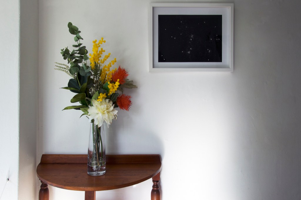Documentation from the contemporary art installation In the Year Twenty Twenty One by Alex Gawronski held at KNULP gallery in Camperdown, Sydney, Australia. A white wall with a black and white photograph in a white frame and an organic abstract painting in brown and gold. A vase with a bunch of colorful artificial flowers on a rounded wooden side table.